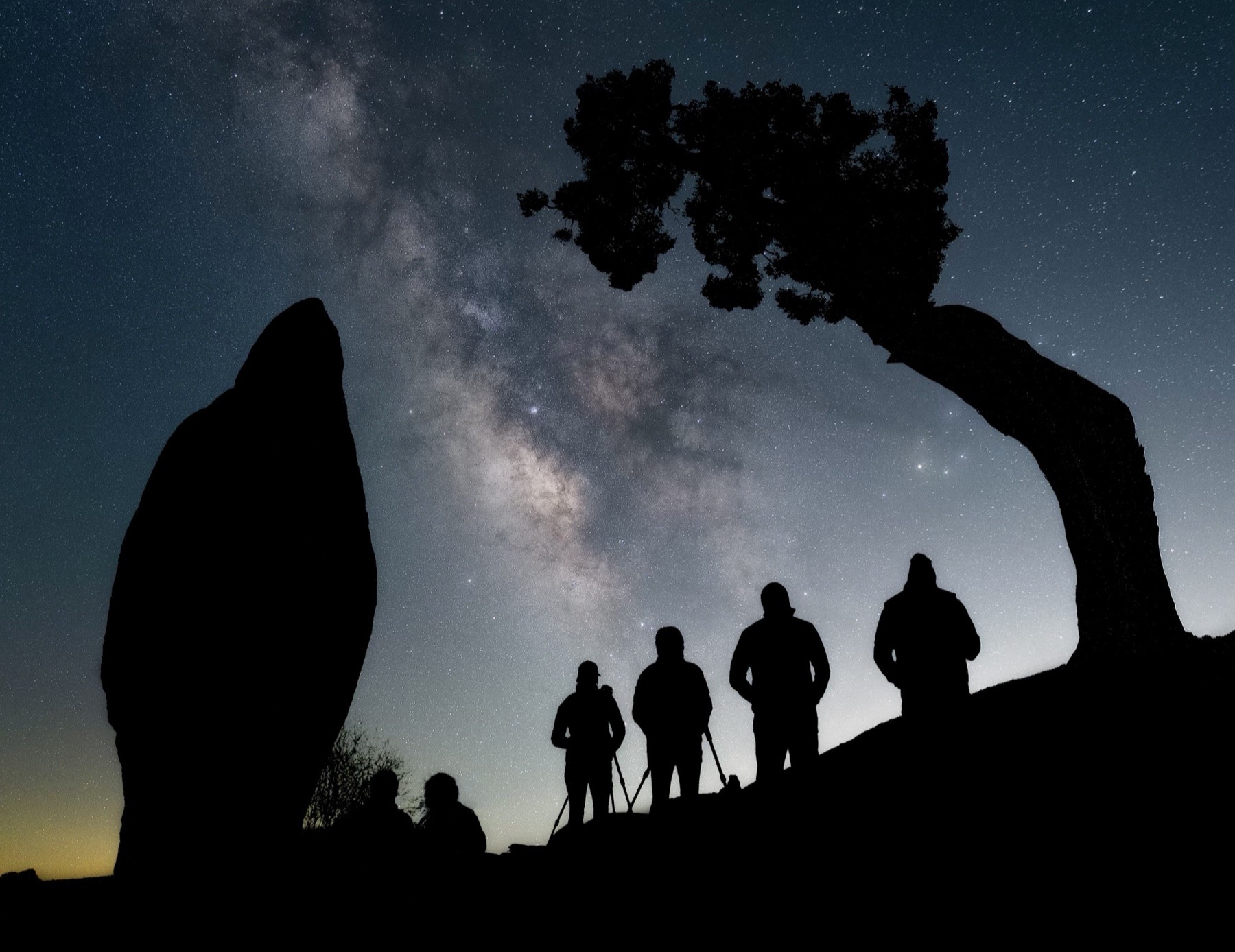 Night photographers silhouetted beneath the Milky Way between desert rock formations and a leaning tree