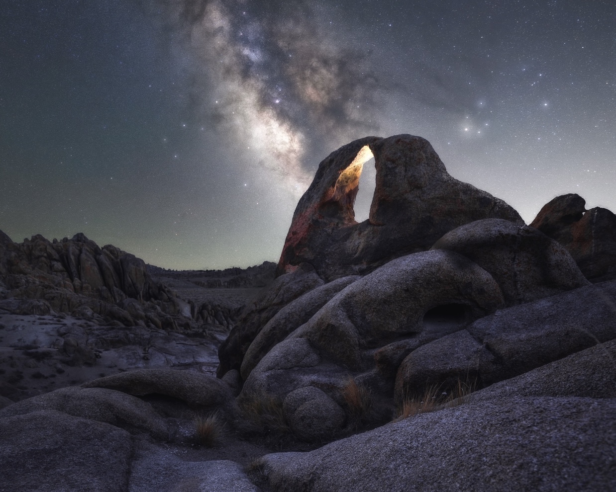 Milky Way core rising above the left side of a small granite arch in Alabama Hills with warm light inside the opening