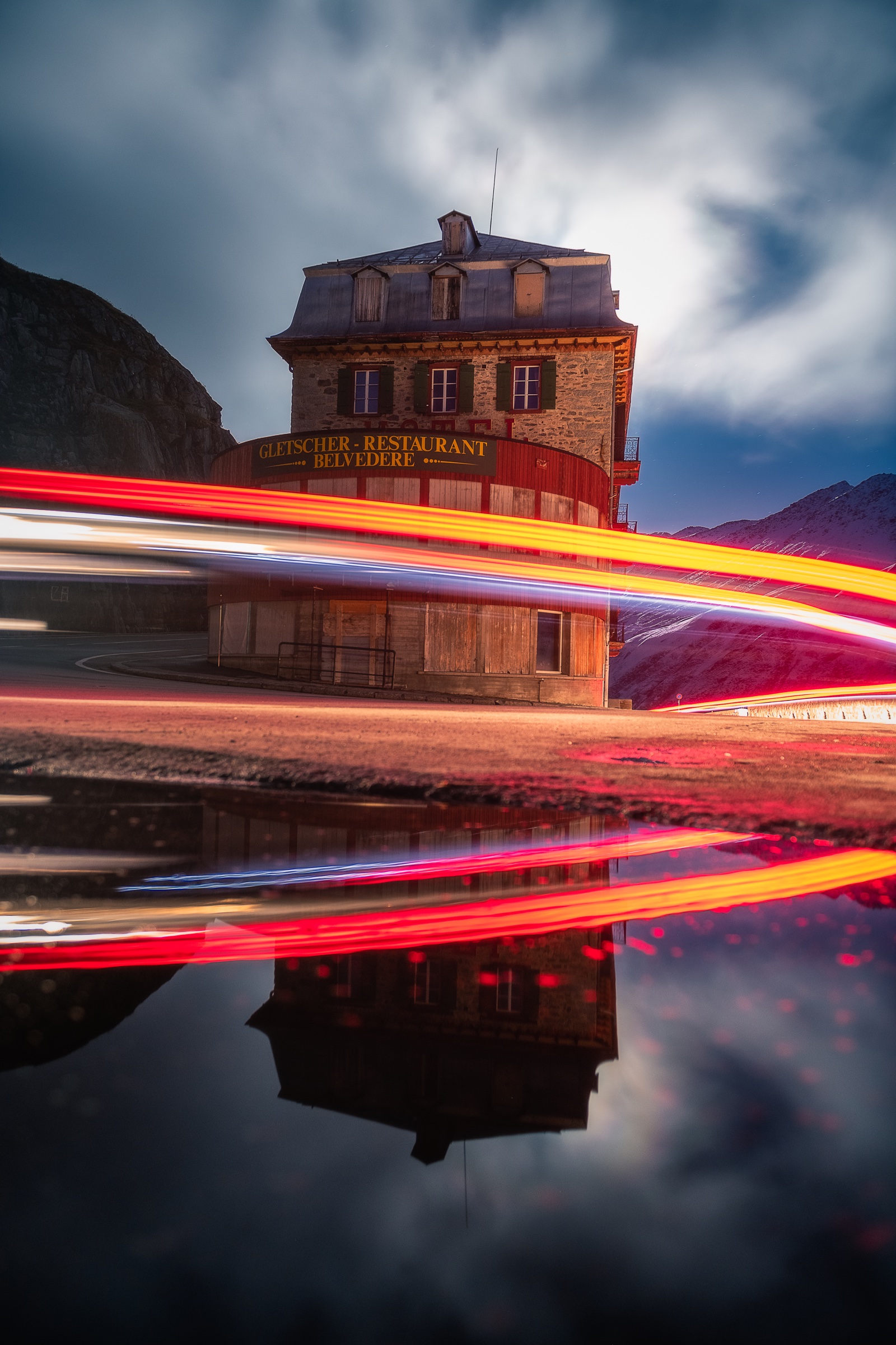 Red and white vehicle light trails crossing a mountain building in Switzerland and reflecting in water