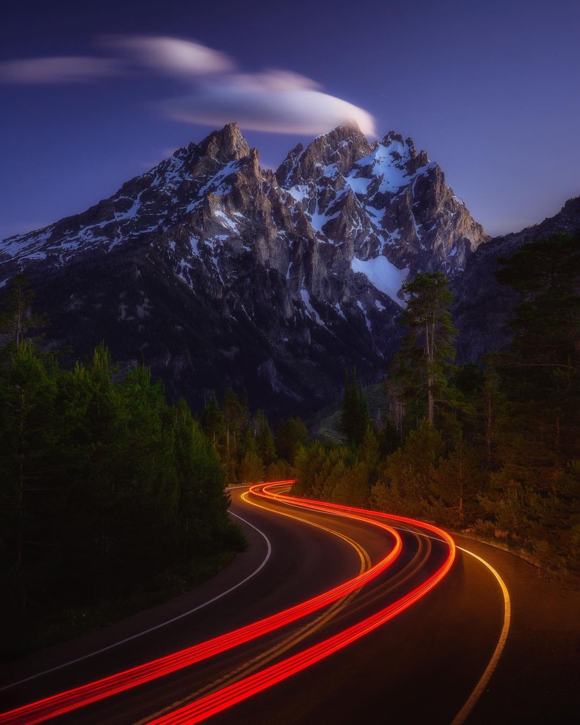 Red vehicle light trails curving beneath snow-covered Teton peaks at blue hour