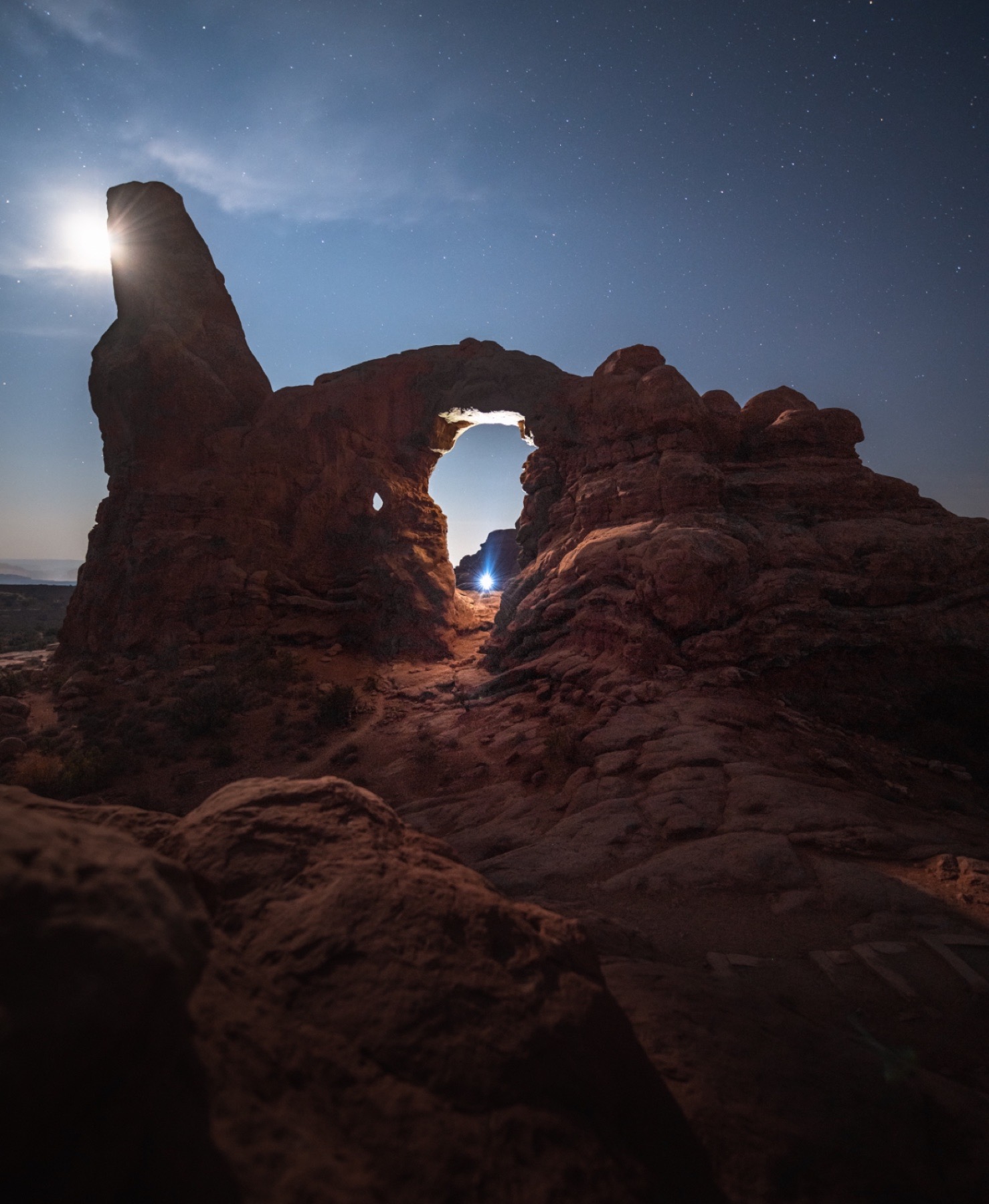 Moonlight breaking around a sandstone spire beside a rock arch under stars