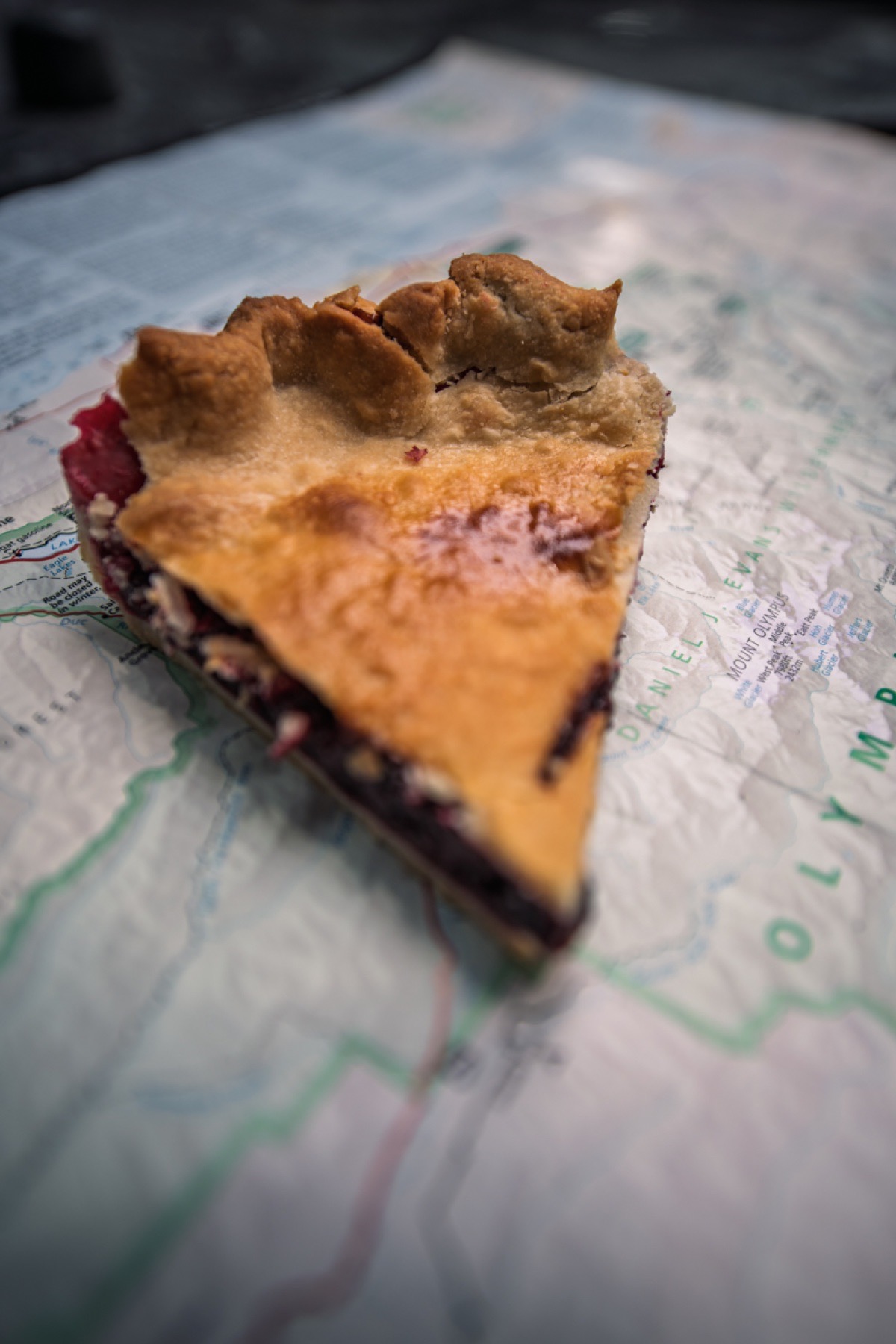 Slice of blackberry pie on a map, with a golden crust and visible berry filling, creating a rustic, adventurous mood.