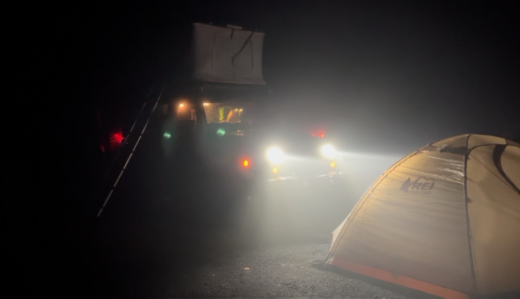 Jeep with bright headlights illuminates a yellow REI tent at night. A ladder is visible, adding a sense of adventure in the dark.