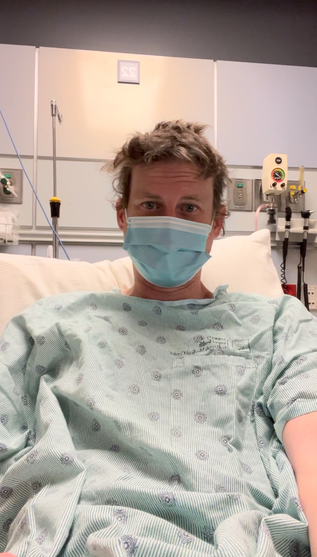 Ryan in a hospital gown and mask sits on a hospital bed.