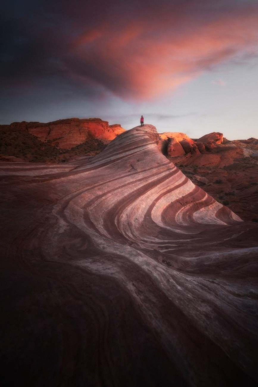 Curved sandstone bands lead toward an isolated person standing on a sunlit ridge beneath a dramatic sky.